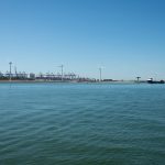 Parque eólico costeiro próximo a um porto, com embarcações e estruturas marítimas ao fundo