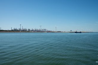 Parque eólico costeiro próximo a um porto, com embarcações e estruturas marítimas ao fundo