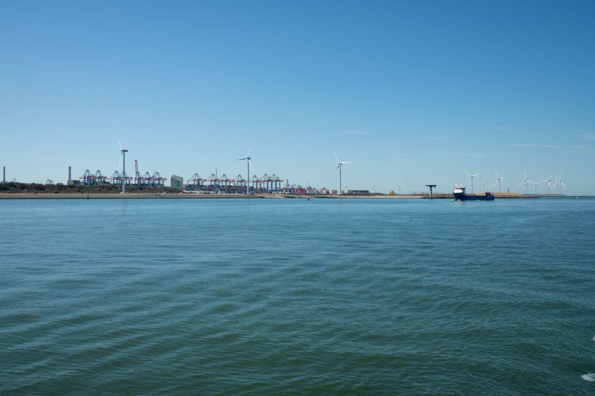 Parque eólico costeiro próximo a um porto, com embarcações e estruturas marítimas ao fundo