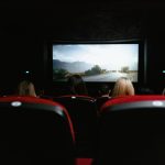 Público visto de costas durante uma sessão de cinema, diante de uma grande tela em uma sala com poltronas vermelhas