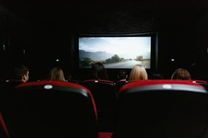 Público visto de costas durante uma sessão de cinema, diante de uma grande tela em uma sala com poltronas vermelhas