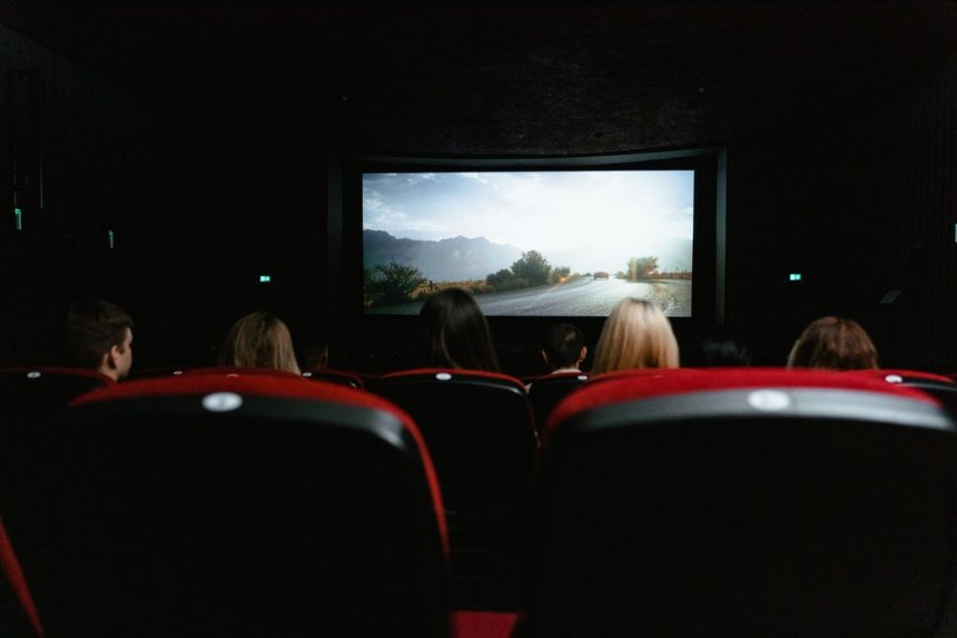 Público visto de costas durante uma sessão de cinema, diante de uma grande tela em uma sala com poltronas vermelhas