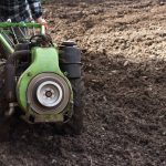 Agricultor opera um pequeno trator em campo recém-arado durante o preparo do solo para o plantio.