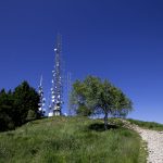 Torres de radiodifusão instaladas em uma colina gramada, sob céu azul, com árvores e caminho rochoso ao redor.