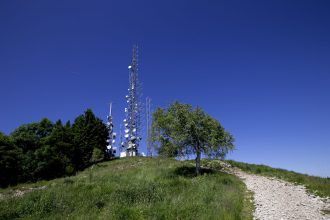 Torres de radiodifusão instaladas em uma colina gramada, sob céu azul, com árvores e caminho rochoso ao redor.