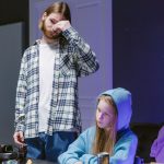 Adolescente joga videogame enquanto um adulto a supervisiona em uma sala.