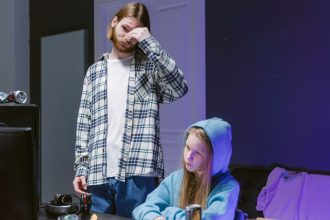 Adolescente joga videogame enquanto um adulto a supervisiona em uma sala.